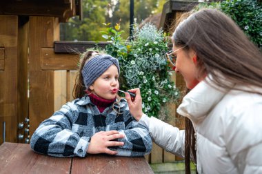 İki kız bahçede bir masada oturuyor ve yaşlı olan da genç olanın dudaklarını boyuyor.