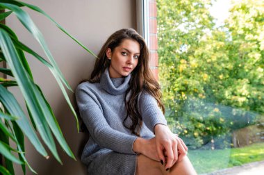 Genç, şehvetli, uzun saçlı esmer kadın evdeki pencere eşiğinde oturuyor.