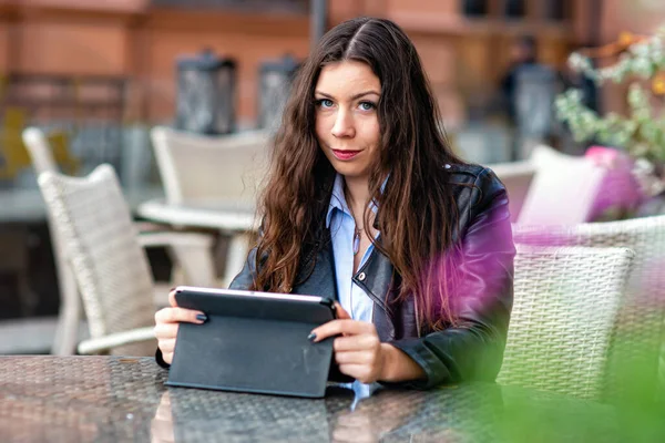 年轻女性坐在街边咖啡店的桌子边 看着相机 一边用平板电脑工作 — 图库照片