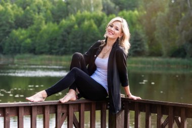 Genç, pozitif, gülümseyen kadın arka plandaki bulanık göl manzarasıyla iskele korkuluklarında oturuyor.