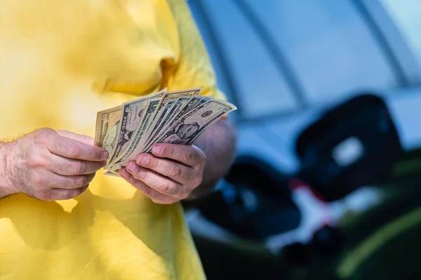 man holding dollar bills on background of car with open gas tank, close-up of hands