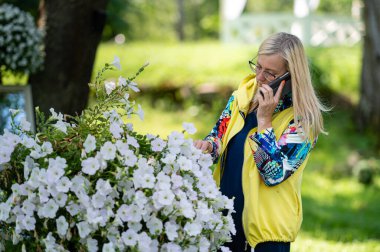 Parlak giyimli, sarışın, son sınıf öğrencisi, bahçede akıllı telefondan konuşan, online iletişim konsepti.