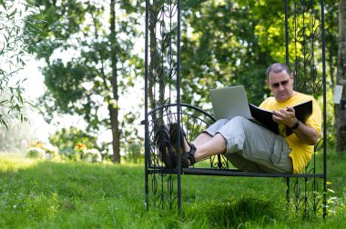 Diz üstü bilgisayarı ve belgesi olan olgun bir adam bahçede çalışıyor, yeşil ofis kavramı..