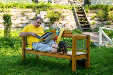 Diz üstü bilgisayarı ve belgesi olan olgun bir adam bahçede çalışıyor, yeşil ofis kavramı..