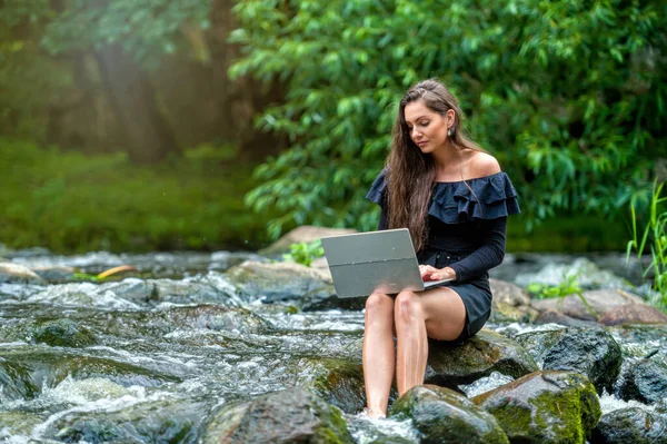 female freelancer sitting on a rock in the river and using a laptop, travel or vacation work concept