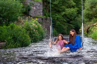 laughing woman and a young girl swing over a fast-flowing river, splashing the water with their feet