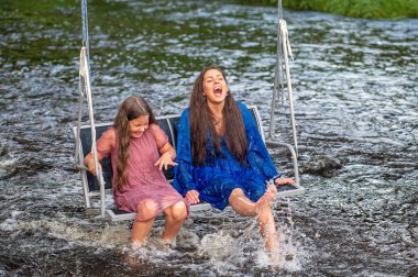 laughing woman and a young girl swing over a fast-flowing river, splashing the water with their feet