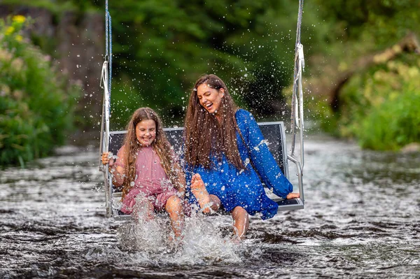 laughing woman and a young girl swing over a fast-flowing river, splashing the water with their feet