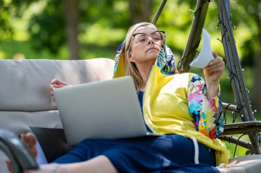Bilgisayarlı ve belgeli olgun bir kadın bahçede sallanan koltukta çalışıyor, yeşil ofis konsepti.