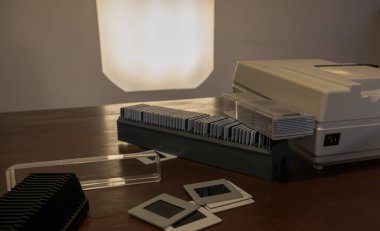 Yandan görünen antika beyaz slayt projektörü, yukarıdan slaytlar saçılmış, projektörün ışığı beyaz duvara yansıtılmış., 