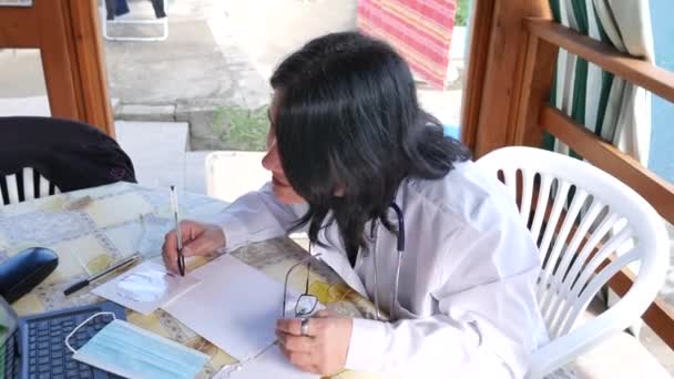 Médecin qui travaille intelligemment depuis la maison, devant l'ordinateur, donnant des conseils et écrivant des recettes par vidéoconférence, sur son patio dans le jardin — Video