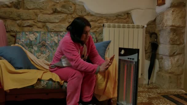 Nette, kühle Frau in rosa Pyjama, die zu Hause auf dem Sofa liegt und ihre Hände und Füße vor einer Elektroheizung wärmt. zitterte in der Wärme der Heimat. — Stockvideo