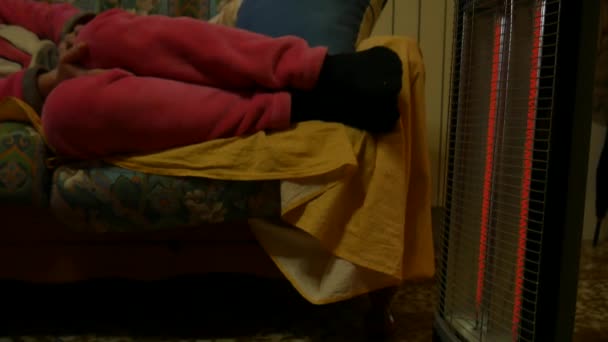 Милая холодная женщина в розовой пижаме, лежащая дома на диване, греет руки и ноги перед электрическим обогревателем. дрожь в тепле дома. — стоковое видео