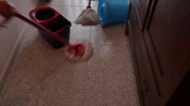 Paspas sıkışıyor, kovaya sıkışıyor, zemini fayanslarla ve arka plandaki renkli perdelerle temizlemeye hazır. İkinci bir paspas ters çevrilmiş..