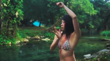 Elegant Asian girl with painted face and body performing an extatic dance in a lush tropical forest near calm pond. 4k