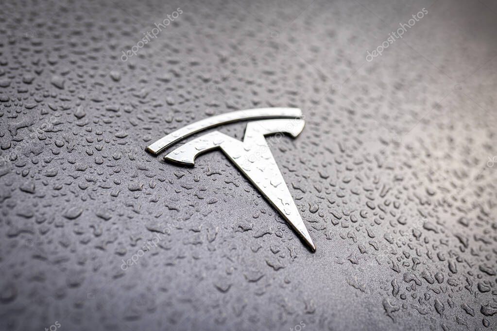 Tesla logo on a grey car with raindrops close-up. Silver badge with water droplets. Tesla Motors, Inc. is an American company that manufactures modern electric cars. Warsaw, Poland - October 23, 2021