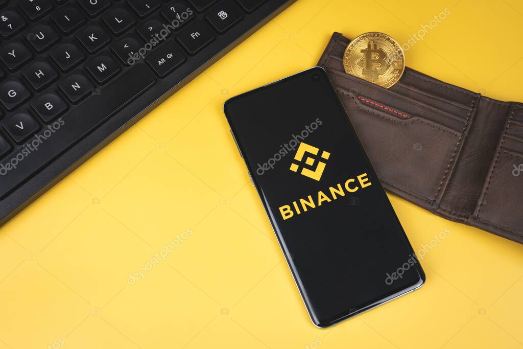 SWANSEA, UK - FEBRUARY 23, 2021: Binance mobile app logo on a smartphone, wallet with gold Bitcoin and keyboard on yellow background. Popular cryptocurrency exchange