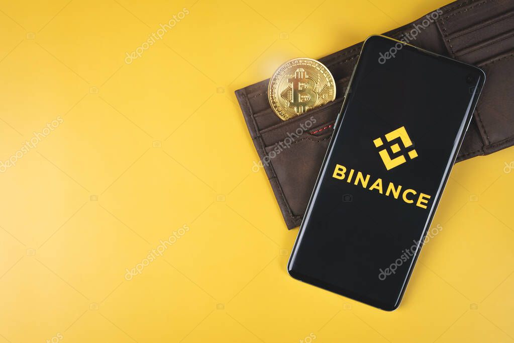 SWANSEA, UK - FEBRUARY 23, 2021: Binance mobile app logo on a smartphone, wallet with gold Bitcoin on yellow background. Popular cryptocurrency exchange