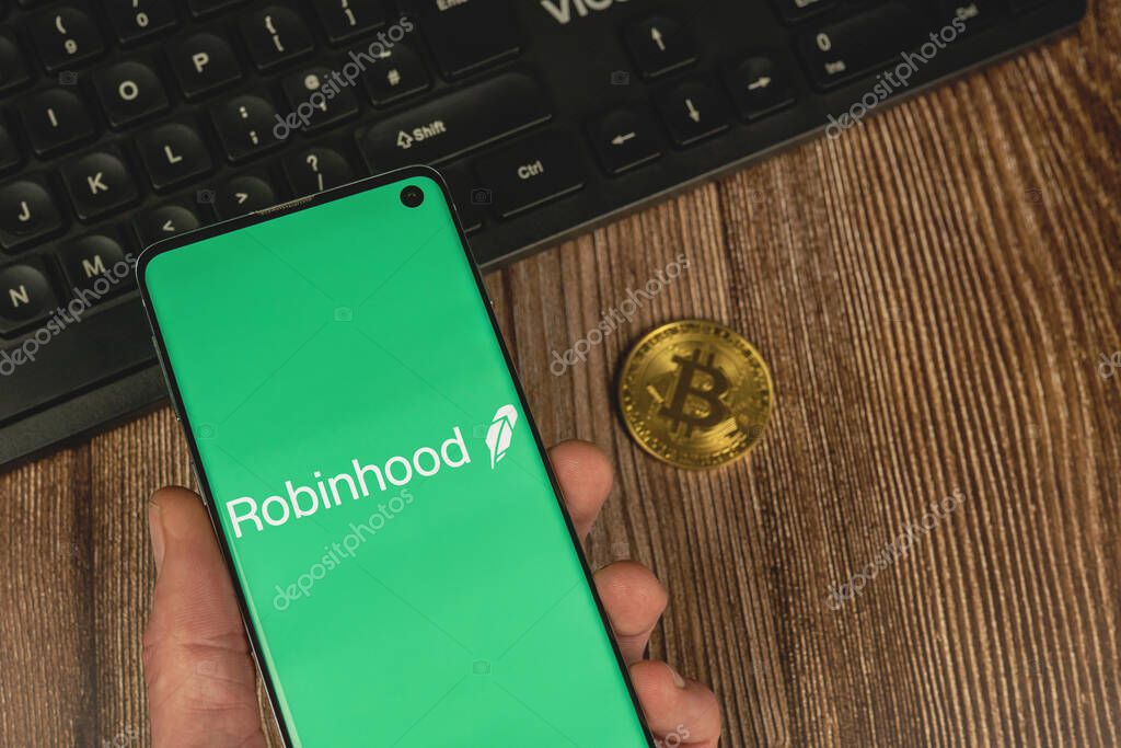 WARSAW, POLAND - FEBRUARY 17, 2021: Man holding a smarphone with Robinhood app green logo with gold Bitcoin and keyboard on wood backround