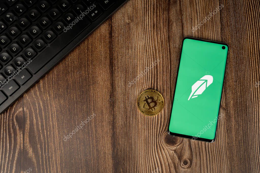 WARSAW, POLAND - FEBRUARY 17, 2021: Robinhood app green logo on smartphone display with gold Bitcoin and keyboard on wood backround