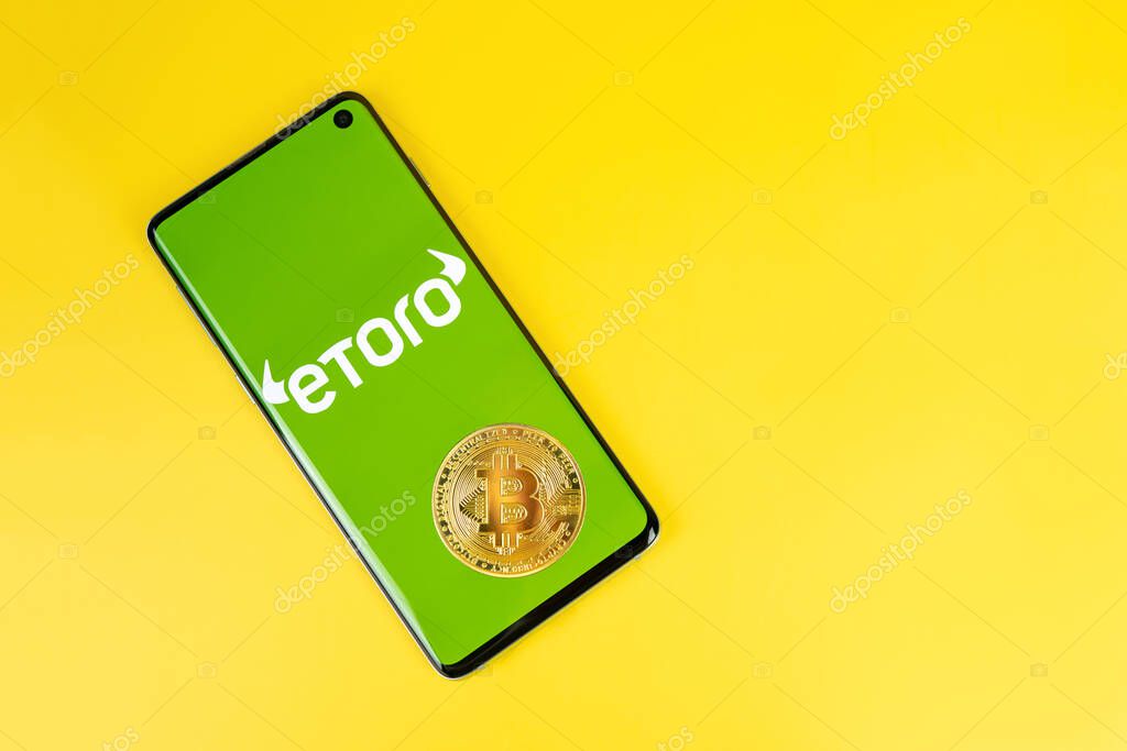 SWANSEA, UK - MAY 4, 2021: Smartphone with Etoro logo with Bitcoin coin on yellow background. Trading platform, investing in stocks and cryptocurrencies