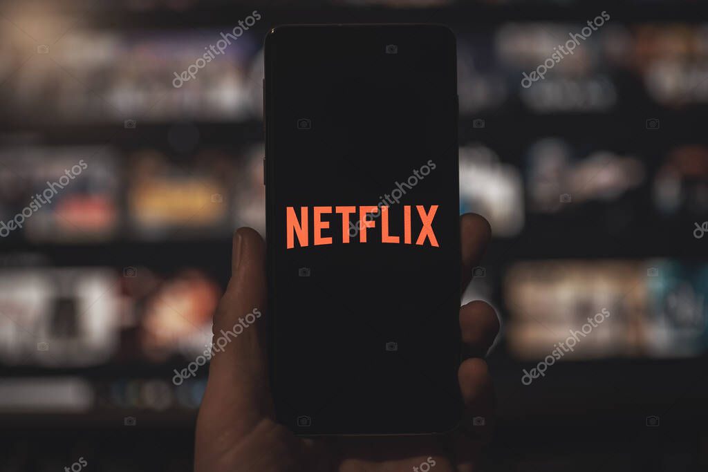 Man holding a smartphone with red Netflix application logo with blurred TV background. Popular streaming service.