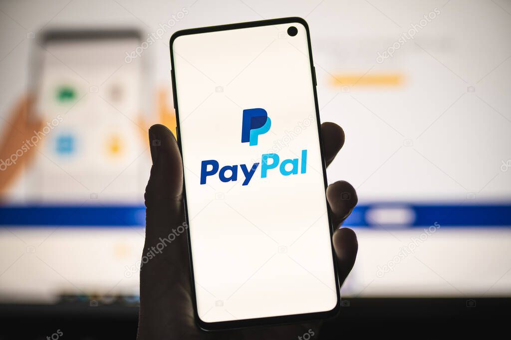Man holding a smartphone with PayPal logo with blurred website as background. Popular online payments system, money transfers