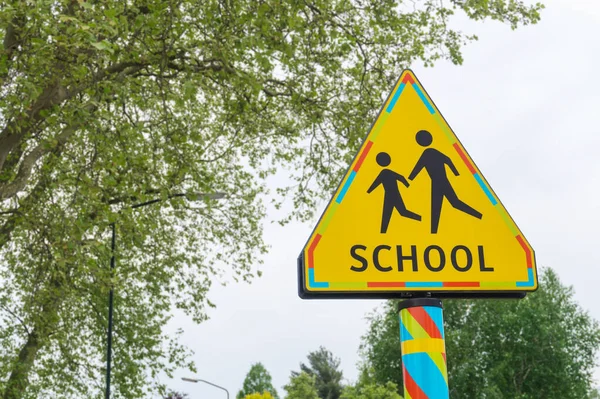 School crossing road sign. Yellow triangle warning for School zone traffic sign in the Netherlands, trees in the background