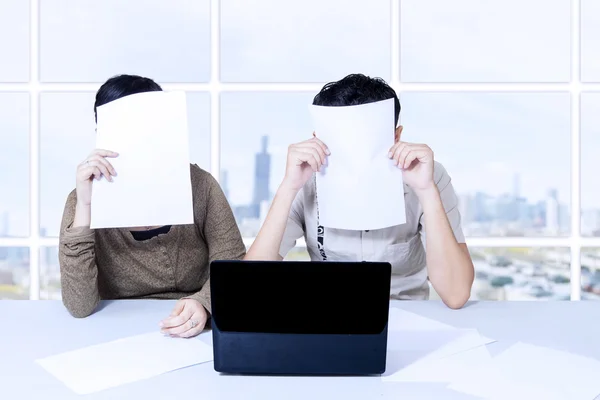 Couple hiding behind papers sitting with laptop — Stock Photo, Image