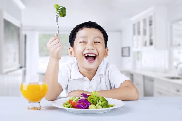 Um menino come salada de legumes na cozinha — Fotografia de Stock