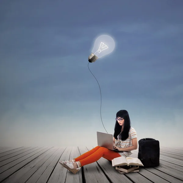 Asian female student typing on laptop under lit bulb — Stock Photo, Image