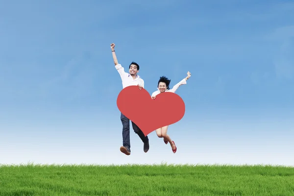 Asiático casal pulando na grama com cartão de coração — Fotografia de Stock