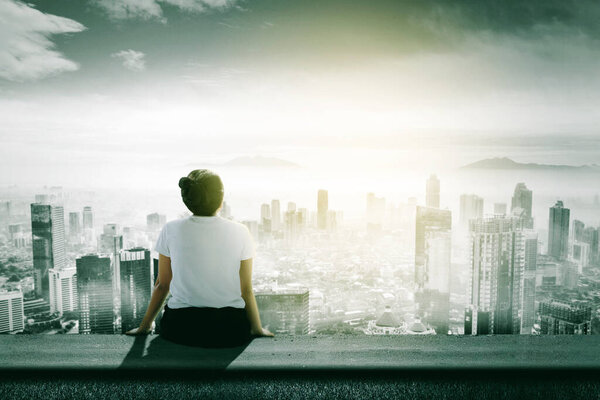 Rear view of young woman looking at misty city while sitting on the apartment roof