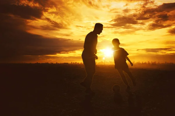 在公园里和儿子踢足球的年轻人的轮廓 具有城市景观背景 黄昏时被枪杀 — 图库照片