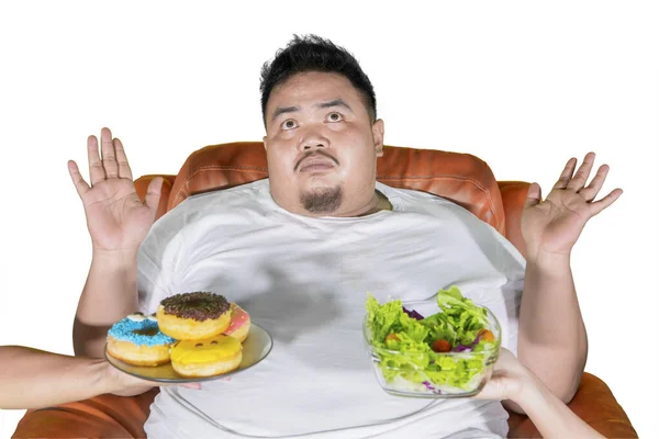 スタジオで野菜サラダやドーナツを選びながら混乱した表情を見せる肥満男 白地に隔離された — ストック写真