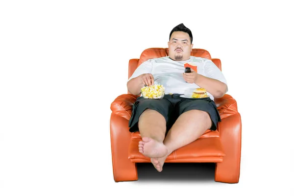 胖年轻人一边吃垃圾食品 一边在演播室看电视 因白人背景而被隔离 — 图库照片