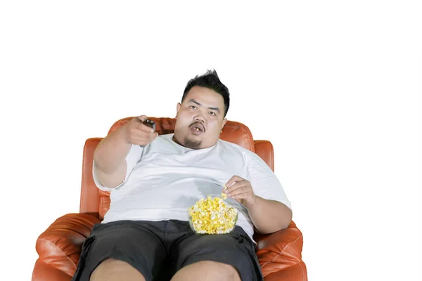 肥胖的年轻人一边看电视 一边坐在扶手椅上 一边吃着一碗爆米花 因白人背景而被隔离 — 图库照片