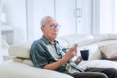 Yaşlı bir adam bir bardak sıcak çay içerken ve evde cep telefonu kullanırken boş vaktinin tadını çıkarıyor.