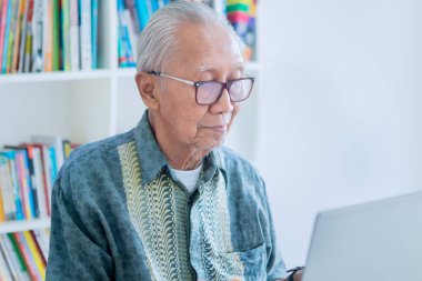 Kütüphanede kitaplığın arkasında otururken dizüstü bilgisayar kullanan yaşlı bir adam