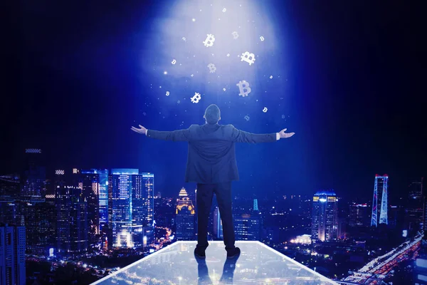 夜市の背景と空から落ちてくるビットコインのシンボルの下に立っている間に手を上げる男性マネージャーの背面図 — ストック写真