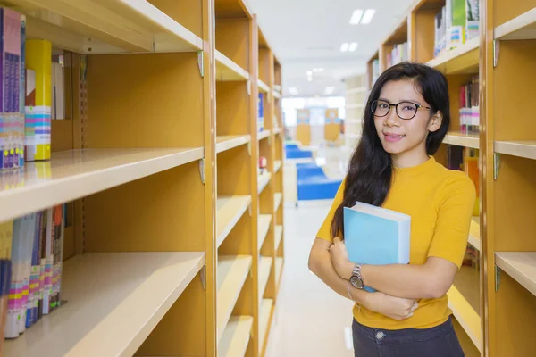 女大学生站在有书架背景的图书馆时拿着书 — 图库照片