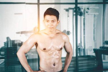 Spor salonunda arka planda duran kaslı genç adam kameraya gülümsüyor.