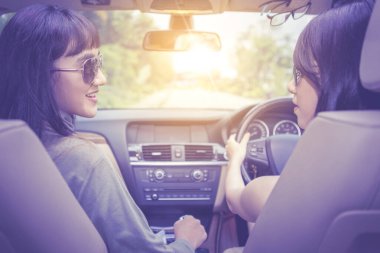 Arka planda iki genç kadın, gün ışığında arabada sohbet ederken araba yolculuğunun keyfini çıkarırken görülüyor.