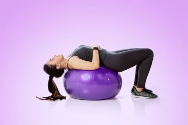 Stüdyoda mor arka planlı bir fitness topuyla karın kaslarını çalıştıran genç bir kadın.