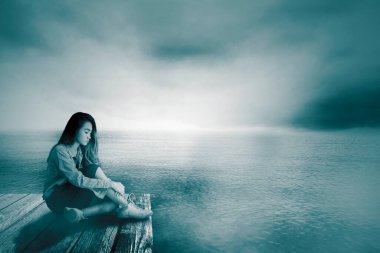 Genç kız, gölün arka planında, ahşap iskelede otururken üzgün görünüyor.