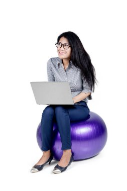 Stüdyoda yoga topunun üzerinde otururken dizüstü bilgisayar kullanan genç bir iş kadını. Beyaz arkaplanda izole