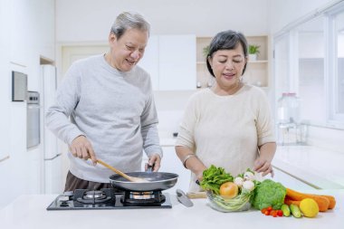 Yaşlı bir adam mutfakta boş vakit geçirirken karısının taze sebzelerle yemek pişirmesine yardım ediyor. Evde vuruldu.