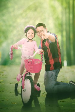 Mutlu baba, kızına parkta bisiklet sürerken kamerayı işaret ediyor.