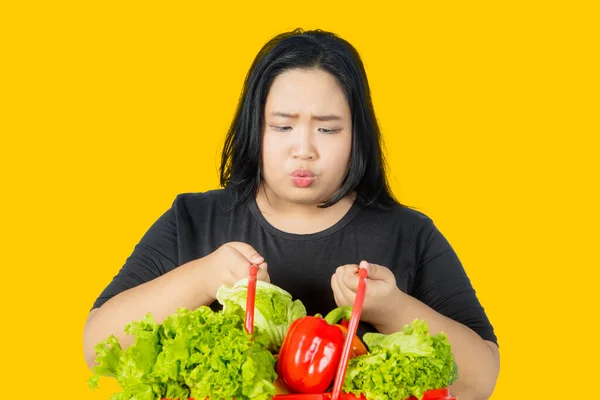 黄色の背景を持つスタジオに立っている間に野菜とショッピングカートを見ていると疑問を感じる太った若い女性 — ストック写真