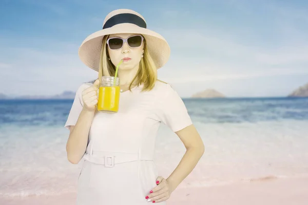 オレンジ色のジュースを楽しみながら熱帯のビーチに立っているいたずらな若い女性 — ストック写真